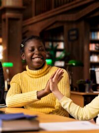 High school students sitting in a library and giving each other a high five working on schoolwork