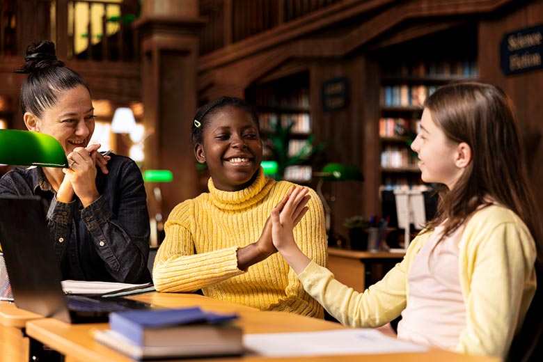 High school students sitting in a library and giving each other a high five working on schoolwork